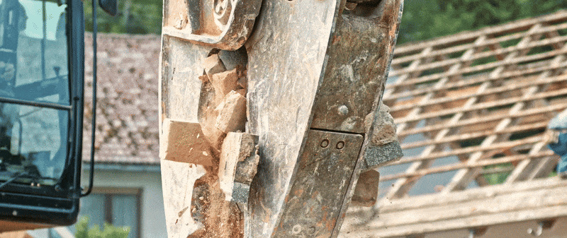 Eine Baggerschaufel voller grober Bauschuttstücke ist im Vordergrund zu sehen. Im Hintergrund erkennt man ein teilweise abgedecktes Gebäude mit Holzdachkonstruktion und grünem Bewuchs. Die Szene vermittelt den Beginn des Recyclingprozesses von Baustoffen.

                          